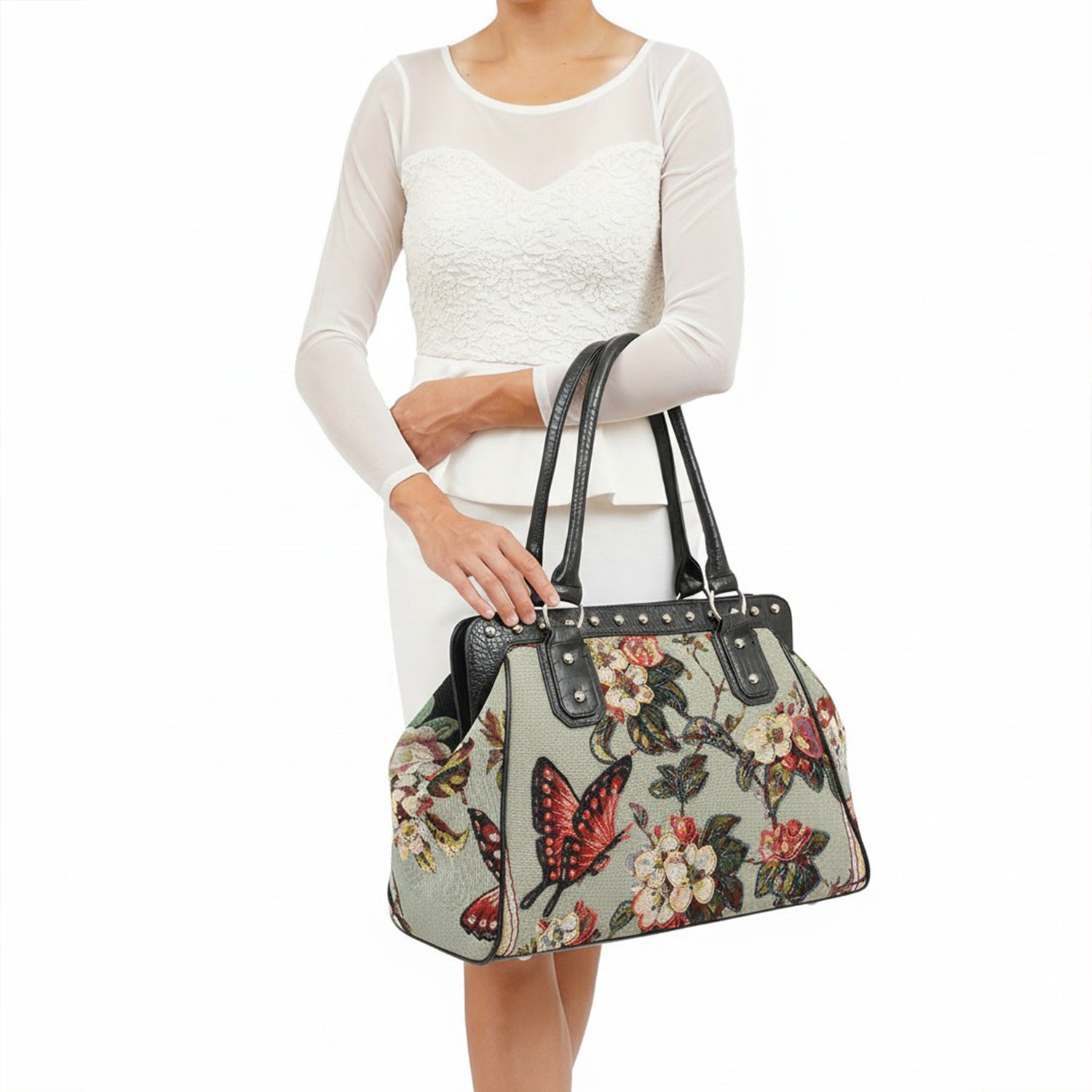 Mellow World floral tapestry handbag with hand-beaded butterfly accents, black vegan‑leather handles, and gold stud detailing, shown held by model in white outfit.